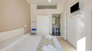 Habitación de hotel con cama, televisor montado en la pared, baño con lavabo y espejo, y una pequeña silla.