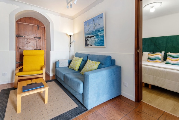 A living room with a blue sofa, a yellow chair, a small wooden table, a painting on the wall, and a door leading to another room.