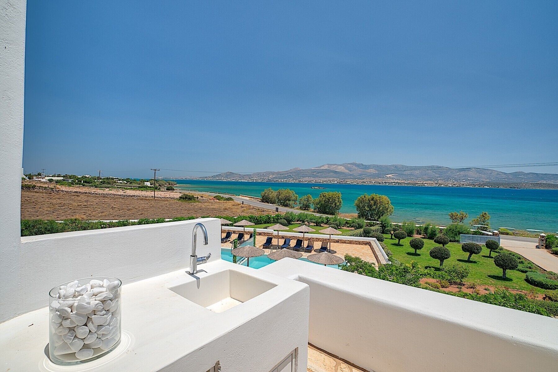 A balcony with a sink, a glass container, and a pool area with lounge chairs and umbrellas.