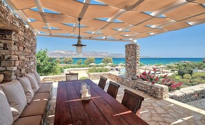A covered outdoor seating area with a table and chairs, stone walls, and a view of the sea. - Villa 'Afroditi B' with Sea View, Wi-Fi and Air Conditioning (Antiparos Town)