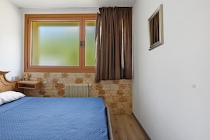 Une chambre à coucher avec un lit, une fenêtre avec des rideaux, une table de chevet en bois et un mur en pierre.
