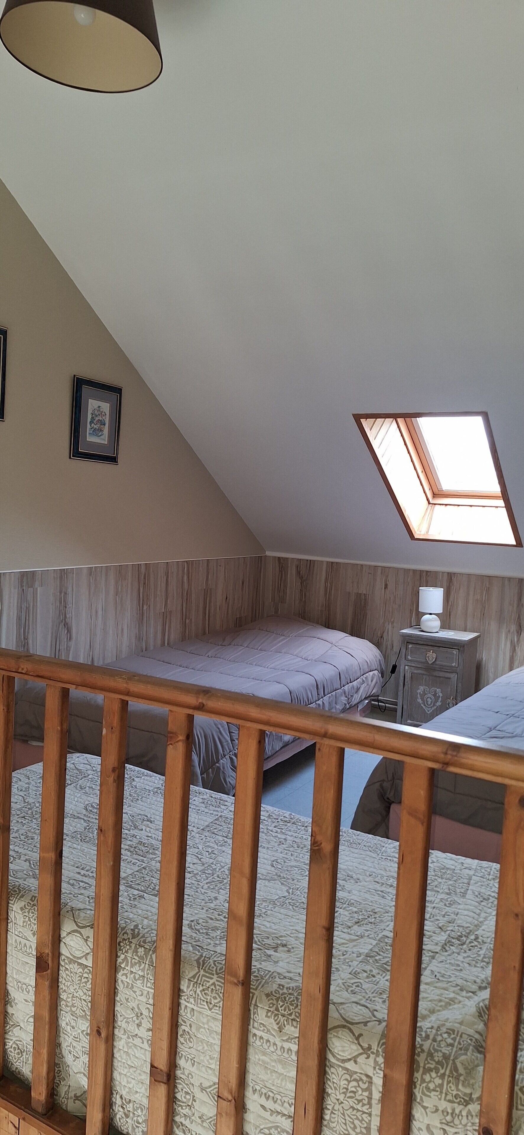 Quarto com teto inclinado, claraboia, duas camas, mesa de cabeceira e corrimão de madeira.