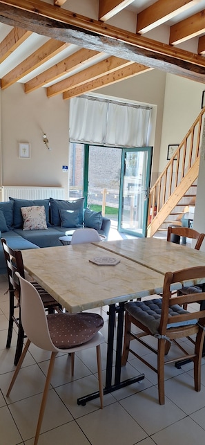 A dining area with a wooden table and chairs, a sofa with cushions, a window with curtains, and a staircase. - Holiday Home 'La Maisonnette Du Meslay' with Private Terrace, Shared Garden & Wi-Fi (Fyé)