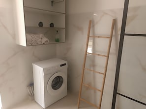 A compact laundry room with a front-loading washing machine, a shelf with folded towels, a small potted plant, and a wooden ladder leaning against the wall. - O'Romarin | Sea & rivers - Chutes du Carbet (Capesterre-Belle-Eau)
