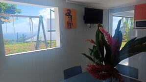 A room with a large window overlooking a landscape, a flat-screen TV mounted on the wall, and a table with a potted plant. - O'Romarin | Sea & rivers - Chutes du Carbet (Capesterre-Belle-Eau)