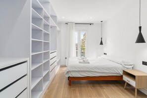 A bedroom with a bed, a wooden nightstand, a white wardrobe, and a view of buildings through a window. - Apartment 'La Siesta 5' with Shared Pool, Wi-Fi and Air Conditioning (Fuengirola)