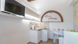 Cozinha compacta com TV integrada, painel de backsplash em pedra, bancada branca e um pequeno espaço de estar com sofá branco.