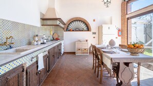 Uma cozinha com painel de azulejos, armários de madeira, bancada branca, luminária pendente e área de jantar com mesa posta para dois.