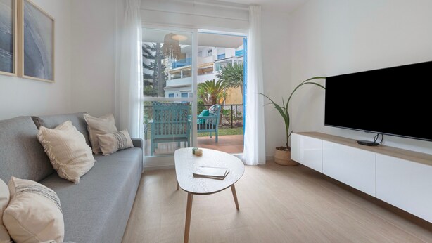 Un soggiorno moderno con divano, tavolino, televisore su mobile bianco e vista su un balcone con mobili da esterno.