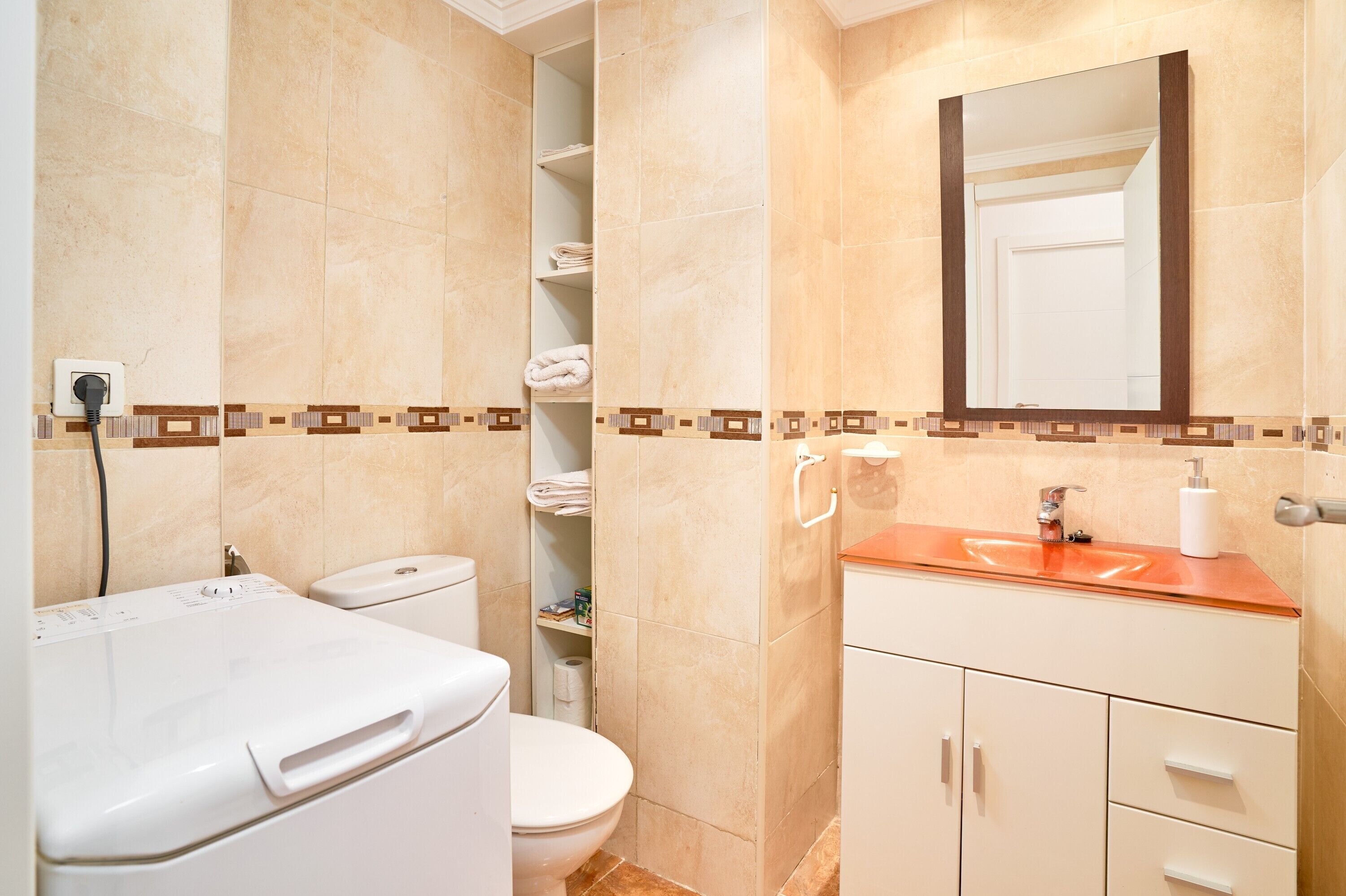 Un baño con inodoro y lavadora blancos, un lavabo con espejo y un estante con toallas.