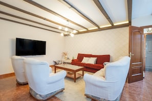 A living room with a flat-screen TV, a red sofa, two white armchairs, a wooden coffee table, and a patterned wall.