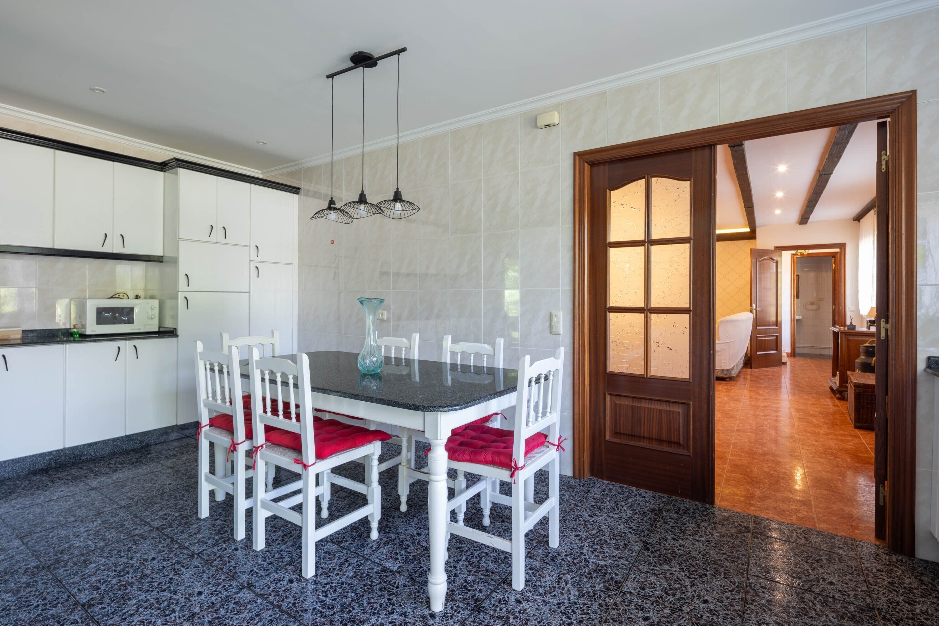 Un coin repas avec une table et des chaises, une cuisine en arrière-plan et une porte en bois donnant sur une autre pièce.