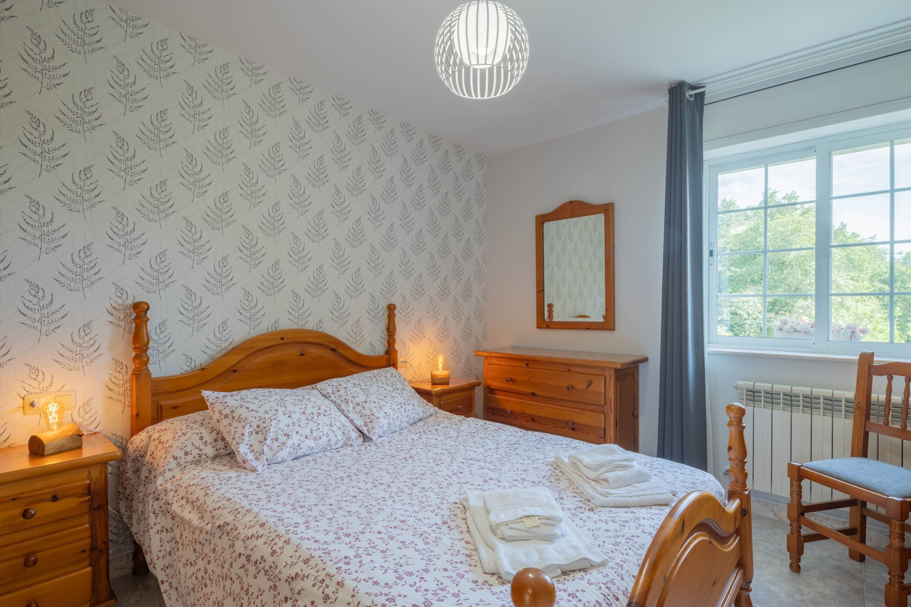 A bedroom with a wooden bed, a nightstand with a lamp, a dresser, a chair, and a window with a view of trees.