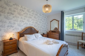 A bedroom with a wooden bed, a nightstand with a lamp, a dresser, a mirror, and a window with a view of trees.