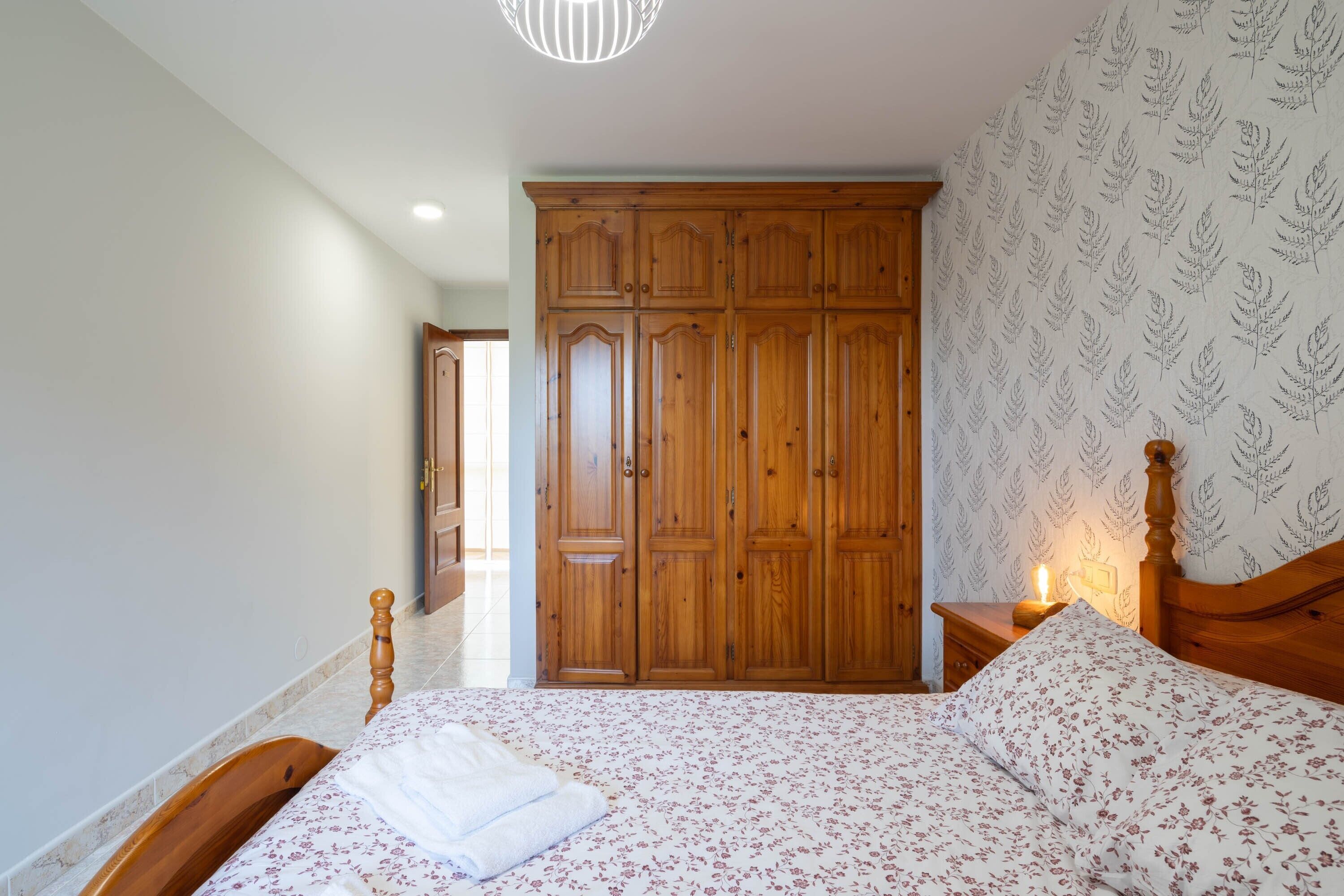 A bedroom with a wooden wardrobe, a bed with floral bedding, and a nightstand with a lamp.