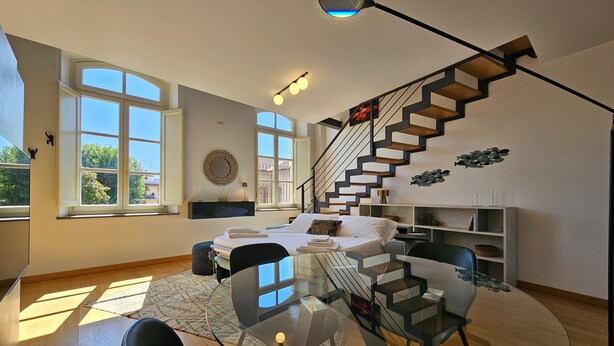 A modern living room with a glass dining table, a sofa, a staircase, and large windows.