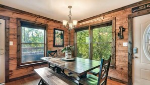 A dining area with a wooden table, chairs, and a chandelier. There is a large window with a view of trees and a door with a sign above it. - 10 mins to Dollywood, Stunning Views, Open Space & HotTub, Pet (Pigeon Forge)
