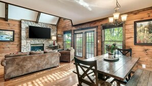 A cozy cabin interior with a stone fireplace, wooden walls, a flat-screen TV, a dining table with chairs, and a large window offering a view of the outdoors. - 10 mins to Dollywood, Stunning Views, Open Space & HotTub, Pet (Pigeon Forge)