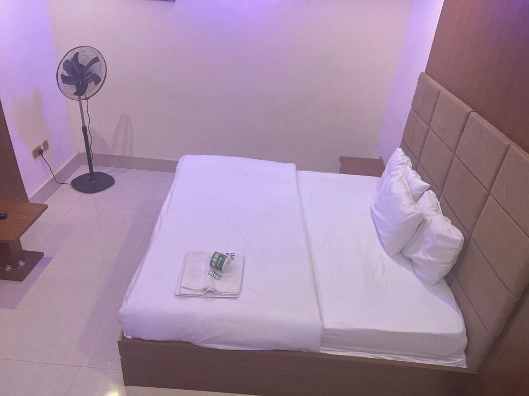 Una cama bien hecha con sábanas blancas, una mesita de noche y un ventilador de pie.