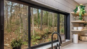 A modern kitchen with a large window overlooking a forest, a built-in sink, and a shelf with kitchenware. - Two Creeks Lodge Modern Creekfront Stay King beds (Cherry Log)