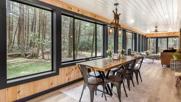 A spacious dining area with a long wooden table, multiple chairs, and large windows offering a view of a forested area. - Two Creeks Lodge Modern Creekfront Stay King beds (Cherry Log)