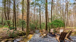 A forest setting with a fire pit and Adirondack chairs. - Two Creeks Lodge Modern Creekfront Stay King beds (Cherry Log)