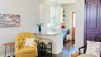 Un salon avec un fauteuil jaune, une table basse noire et une chaise en bois avec un coussin. On aperçoit, au fond, une cuisine avec des meubles blancs et une porte violette.