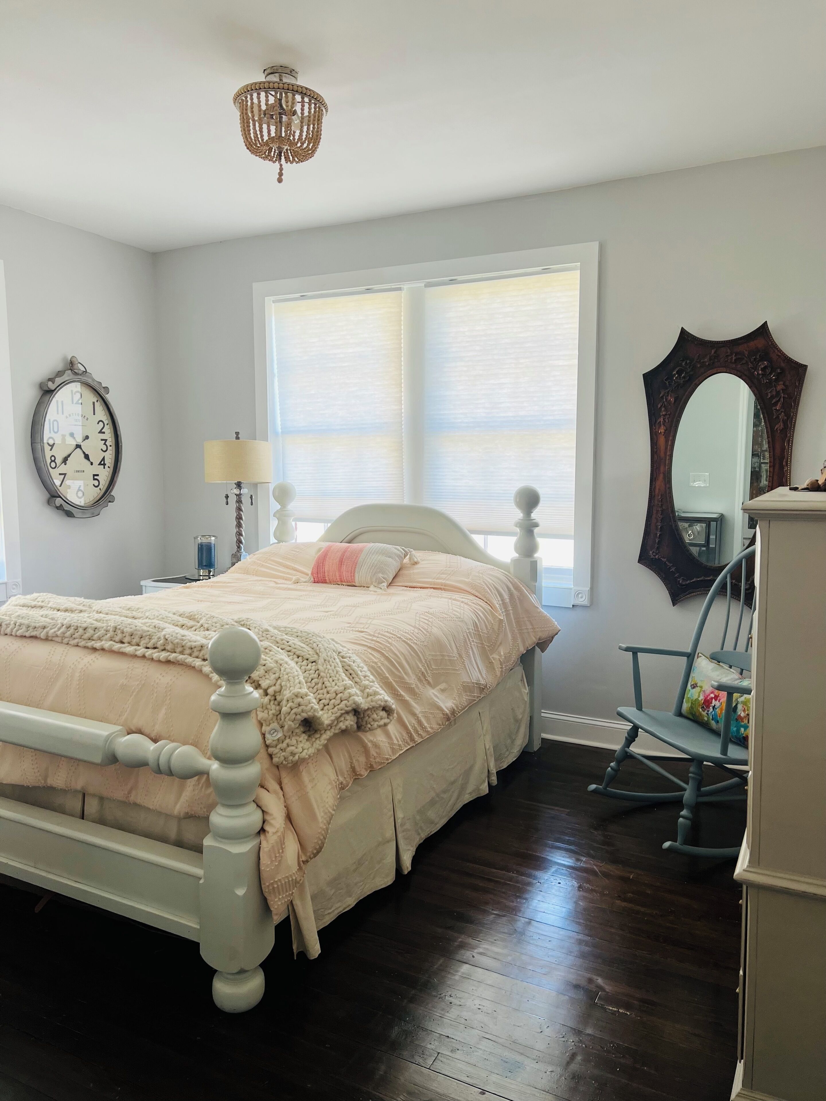 Une chambre à coucher comprenant un lit, une commode, un fauteuil à bascule, un lustre, une fenêtre avec des stores et une horloge murale.