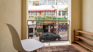 A corner of a room with a white chair, a small table, and a view of a sushi restaurant with a car parked outside. - Sesimbra Beach Apartment by GT House (Sesimbra)