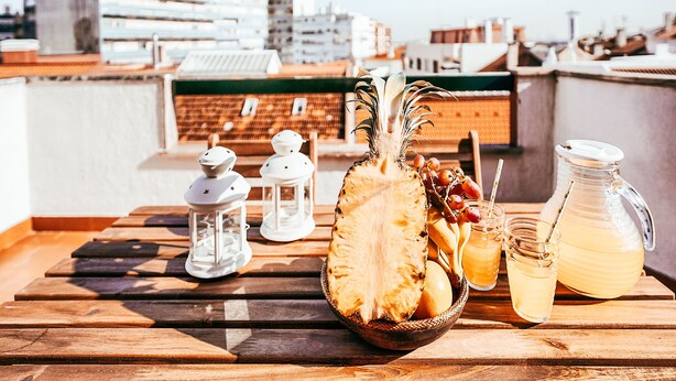 A wooden table with a pineapple, fruits, drinks, and a lantern on a rooftop with city buildings in the background. - Amadora Terrace View By Gt House (Amadora)