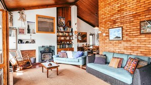 Una sala de estar acogedora con techo de madera, pared de ladrillo, sofá azul, mecedora de madera y una estantería llena de libros y objetos decorativos.