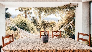 Una mesa de comedor con mantel estampado, rodeada de sillas de madera, con una hamaca y diversas plantas al fondo.