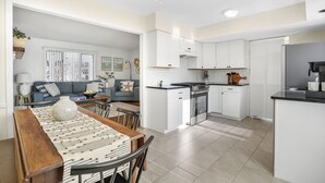 Una cocina moderna con armarios blancos, un comedor con mesa y sillas de madera, y una sala de estar con sofá gris y cojines decorativos.
