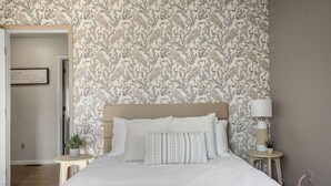 Un dormitorio con papel tapiz estampado, una cama con ropa de cama blanca, una mesita de noche con lámpara y una planta pequeña.