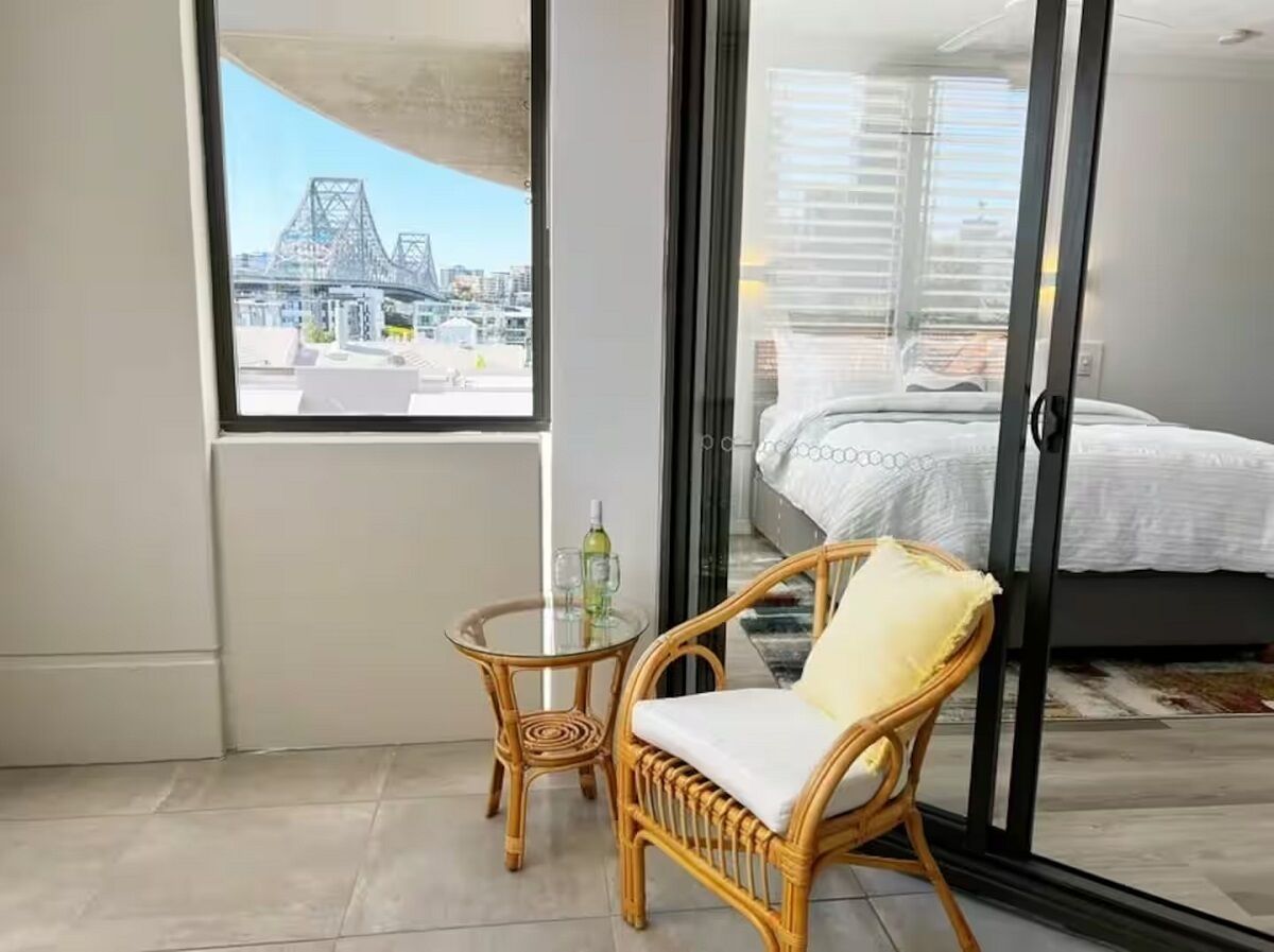 A balcony with a wicker chair and a small table, a bed with a white bedspread, and a view of a bridge and buildings.