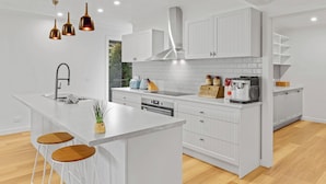 Una cocina moderna con armarios blancos, una isla central con taburetes, horno empotrado y cafetera.