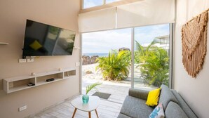 Ein modernes Wohnzimmer mit großem Fernseher, einem grauen Sofa, einem kleinen weißen Tisch und Blick auf einen Strand sowie Palmen durch große Glasfenstertüren.