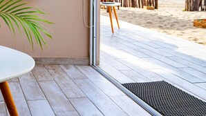 Ein Zimmer mit Holzboden, einem weißen Tisch, einer Topfpflanze, einem an der Wand montierten Fernseher und Blick auf den Strand durch eine offene Tür.