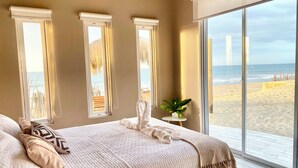 Ein Schlafzimmer mit einem großen Bett, drei Fenstern mit Vorhängen, einem kleinen Tisch mit Pflanze und Blick auf Strand und Meer.