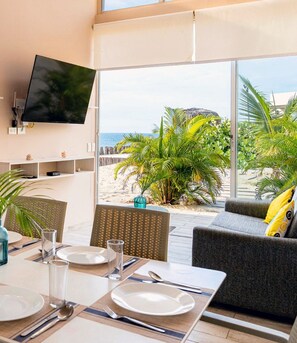 A room with a dining area, a flat-screen TV mounted on the wall, and a large window overlooking a beach with palm trees. - A charming beachfront bungalow overlooking the warm, calm sea. (Canoas)