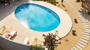 Ein Swimmingpool mit Meerblick, umgeben von einer Sandfläche mit Liegestühlen und einer überdachten Sitzgelegenheit.