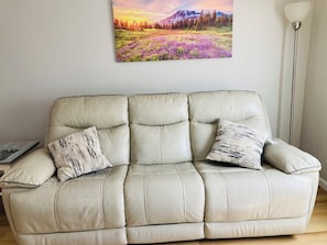 Ein beiges Ledersofa mit zwei gemusterten Kissen, ein gerahmtes Landschaftsbild darüber und eine Stehlampe zur Rechten.