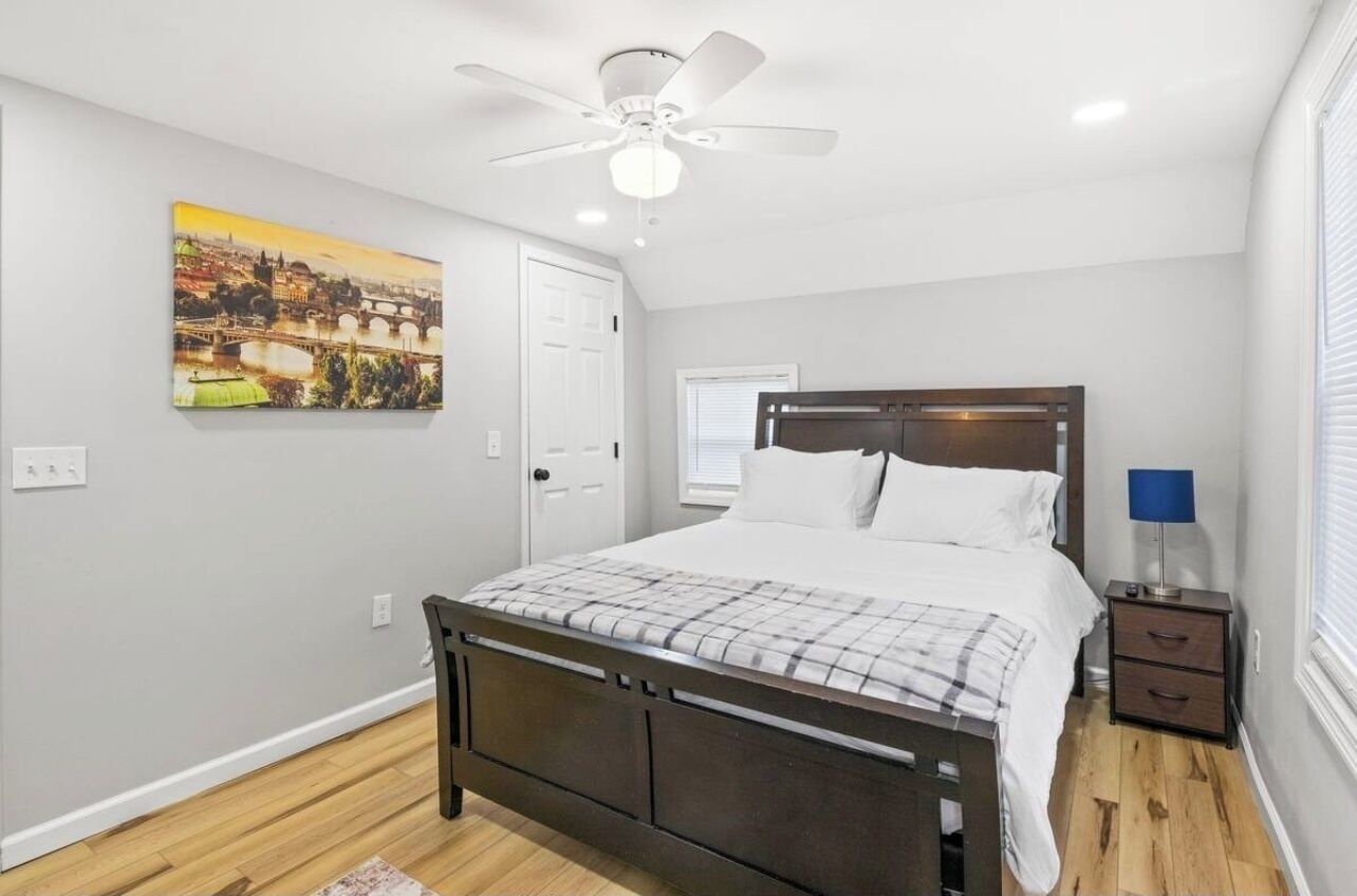 A bedroom with a bed, a ceiling fan, a nightstand, and a painting on the wall.