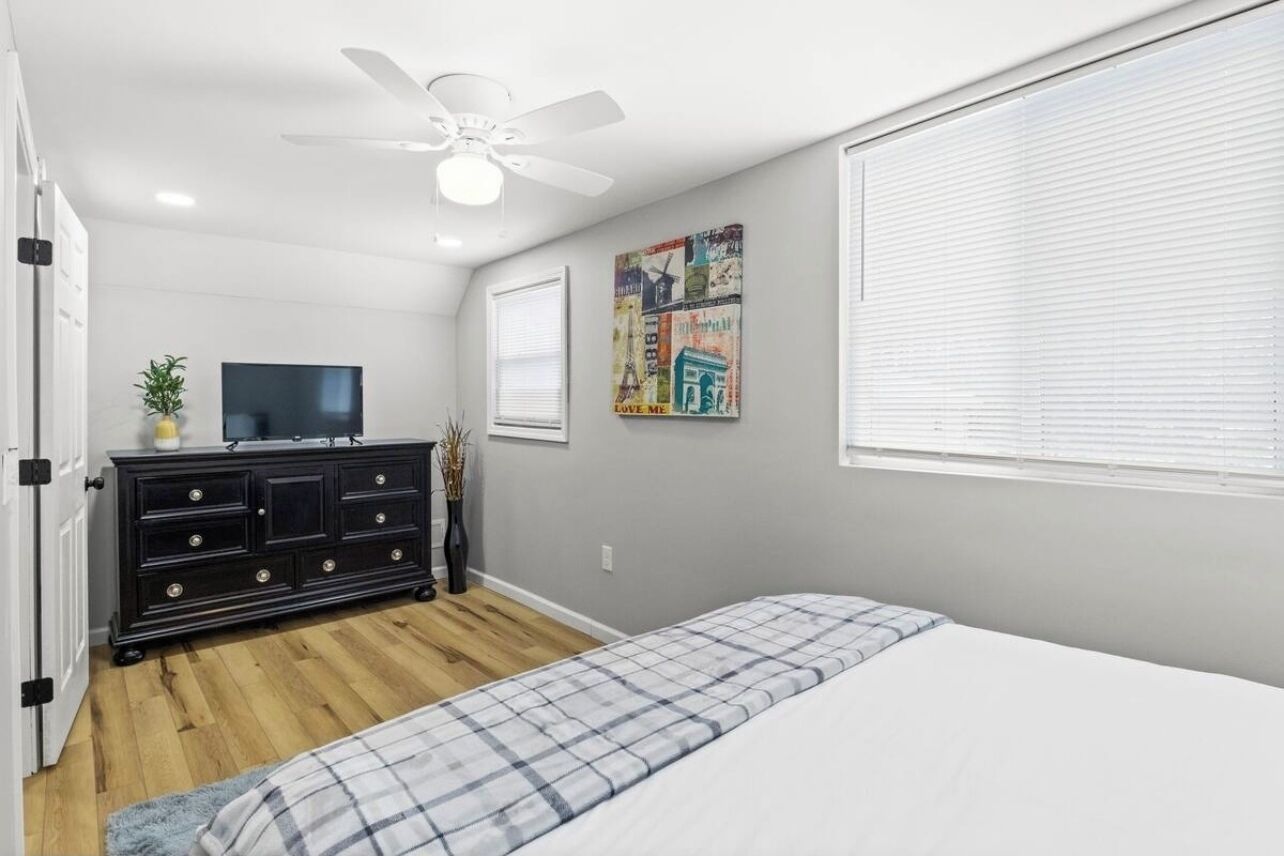 Une chambre à coucher comprenant un lit, une commode, un téléviseur, un ventilateur de plafond et une fenêtre avec des stores.