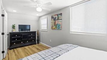 Une chambre à coucher comprenant un lit, une commode, un téléviseur, un ventilateur de plafond et une fenêtre avec des stores.
