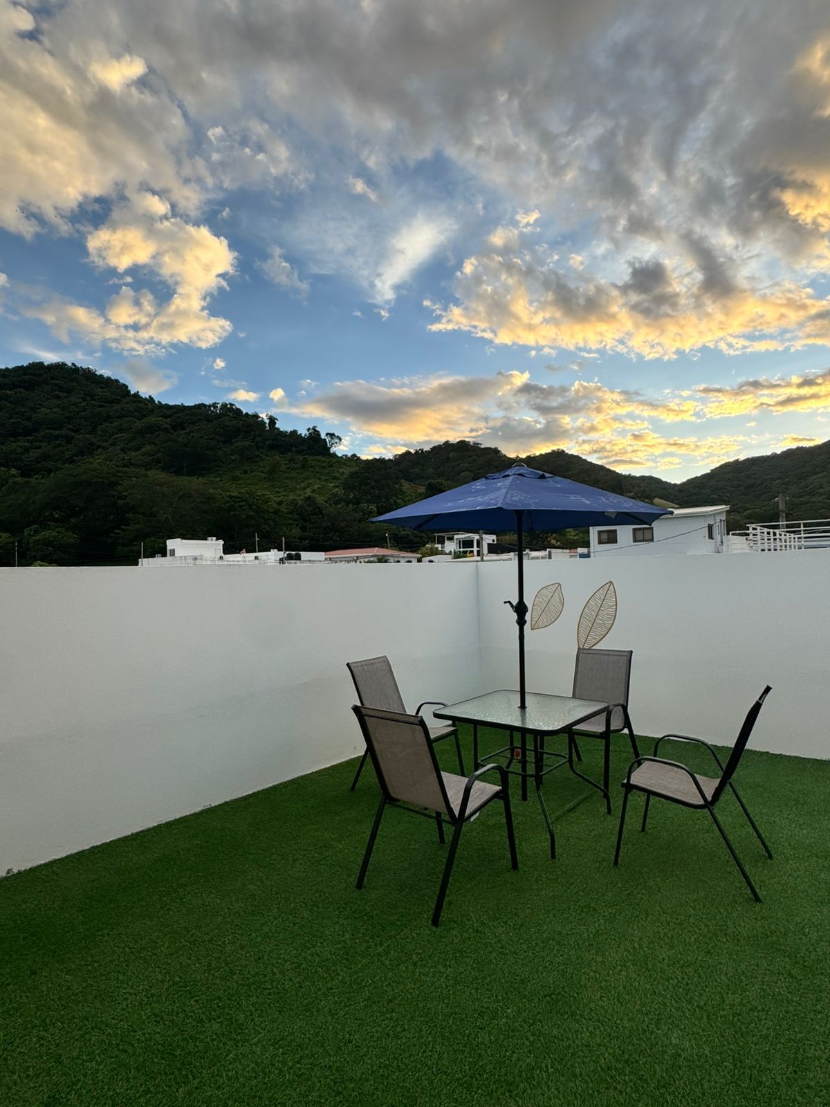 Una terraza en la azotea con una mesa y sillas, una sombrilla azul y vistas a una colina boscosa.