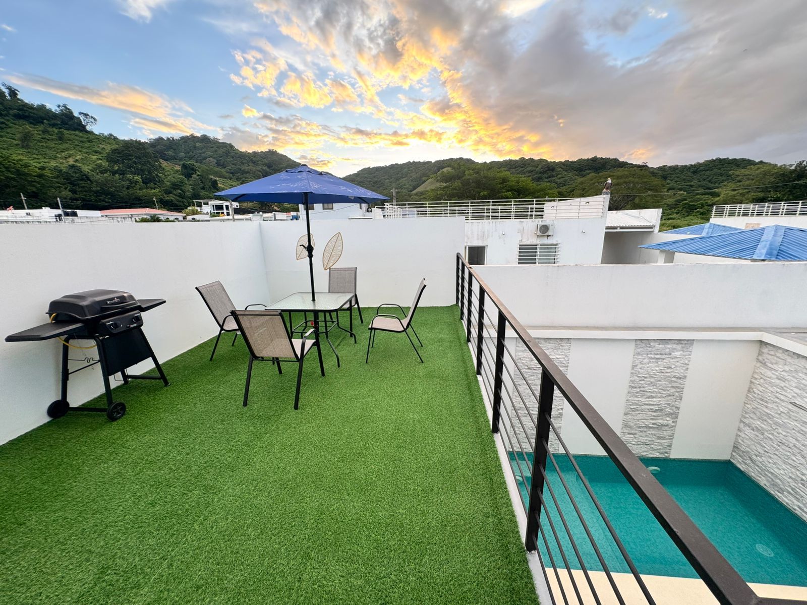 Una terraza en la azotea con césped artificial, una parrilla, una mesa con sillas y una piscina.