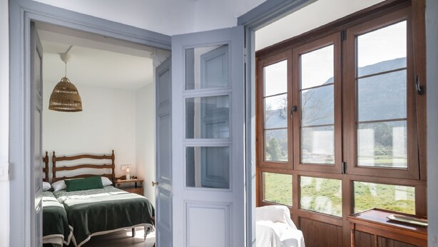 A room with a bed, a nightstand, a hanging light, and a large window with a view of greenery.