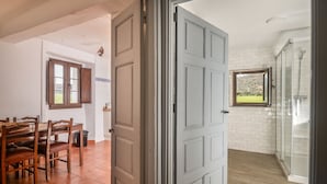 An open door leading to a room with a shower, a dining area with a table and chairs, and a window with a view of greenery.