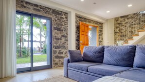 A modern living room with a stone wall, a blue sofa, and a large sliding glass door leading to a garden. - Ricardo House, a Home in Madeira (Calheta)